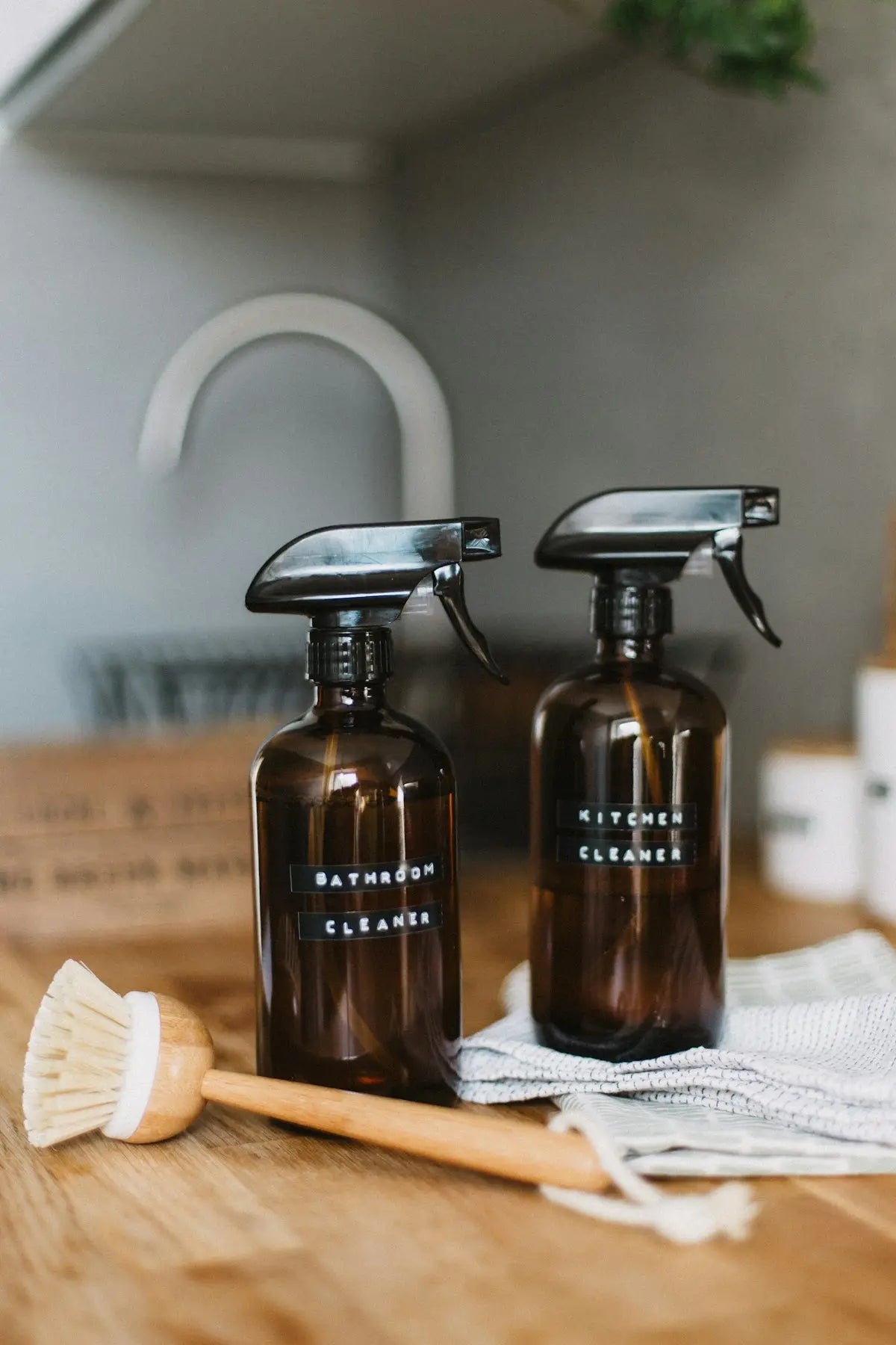 Two amber glass spray bottles labeled bathroom cleaner and kitchen cleanser on wooden countertop for home essentials comparison.