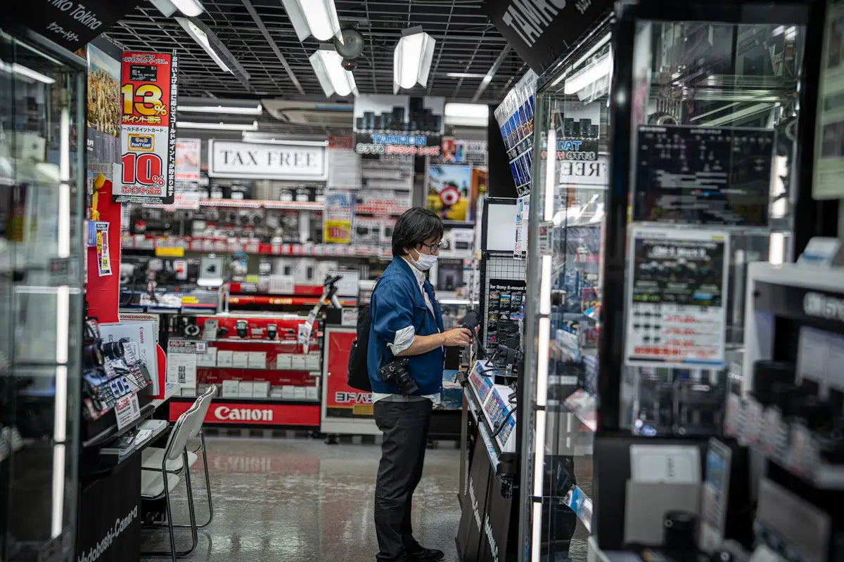 A person wearing a blue jacket and black pants holds a camera in an electronics store aisle for confident tech shopping.