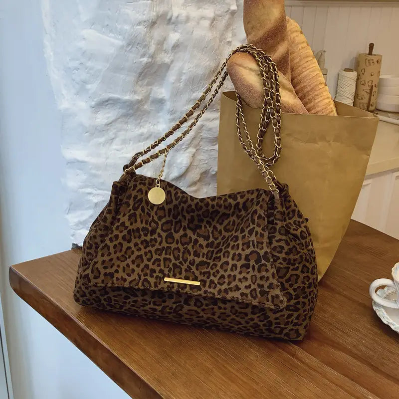 Brown leopard-print crossbody bag made of suede with a gold-tone chain strap and circular metal charm.