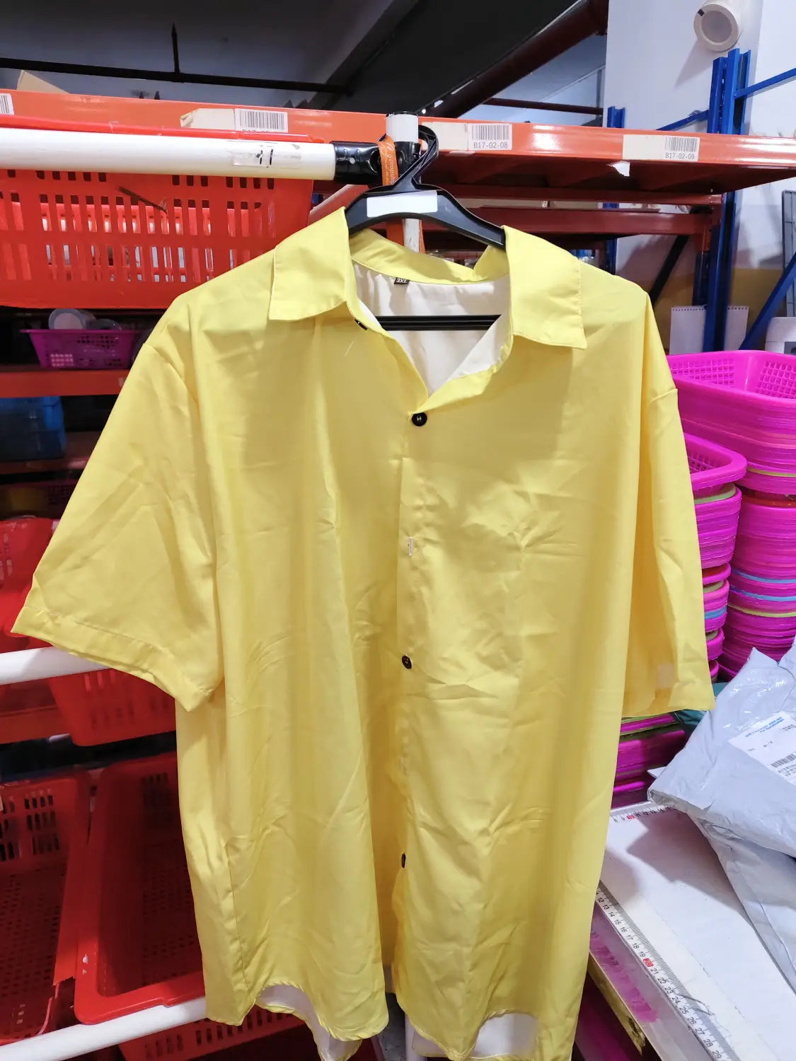 Ein hellgelbes Herren-Muscle-Fit-Poloshirt mit kurzen Ärmeln, schwarzen Knöpfen und einem klassischen Kragen, das an einem Kleiderbügel in einer Lagerumgebung hängt.
