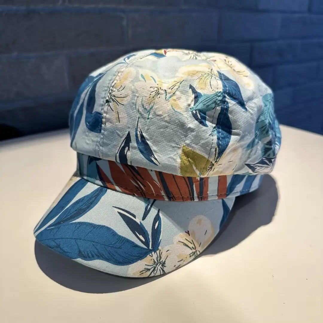 A light blue floral newsboy cap for women featuring bold navy and cream blossoms with a rust and teal striped brim made of fabric.