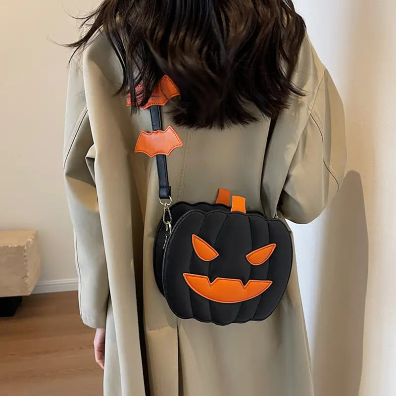 Black pumpkin-shaped crossbody bag with orange stitched facial features and matching orange strap accent featuring a detachable bat charm.