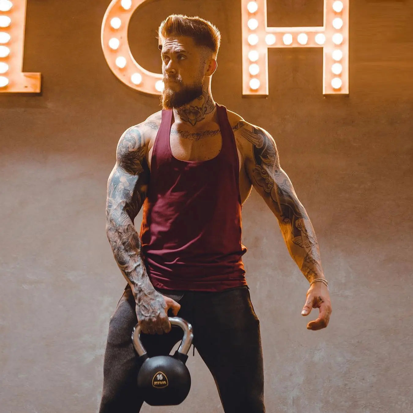 Mens go hard racerback athletic tank top displayed beside a black 16-pound kettlebell with silver handle and gold logo on the base.