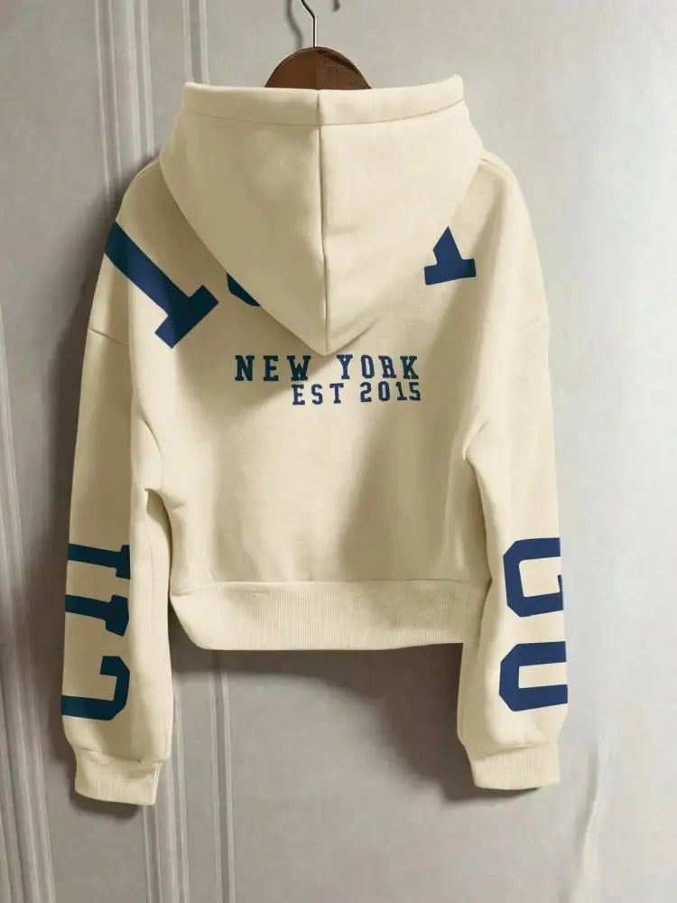 Sweat-shirt zippé court couleur écrue, présenté depuis le dos, avec une impression graphique bleue audacieuse et des lettrages « New York Est 2015 ».