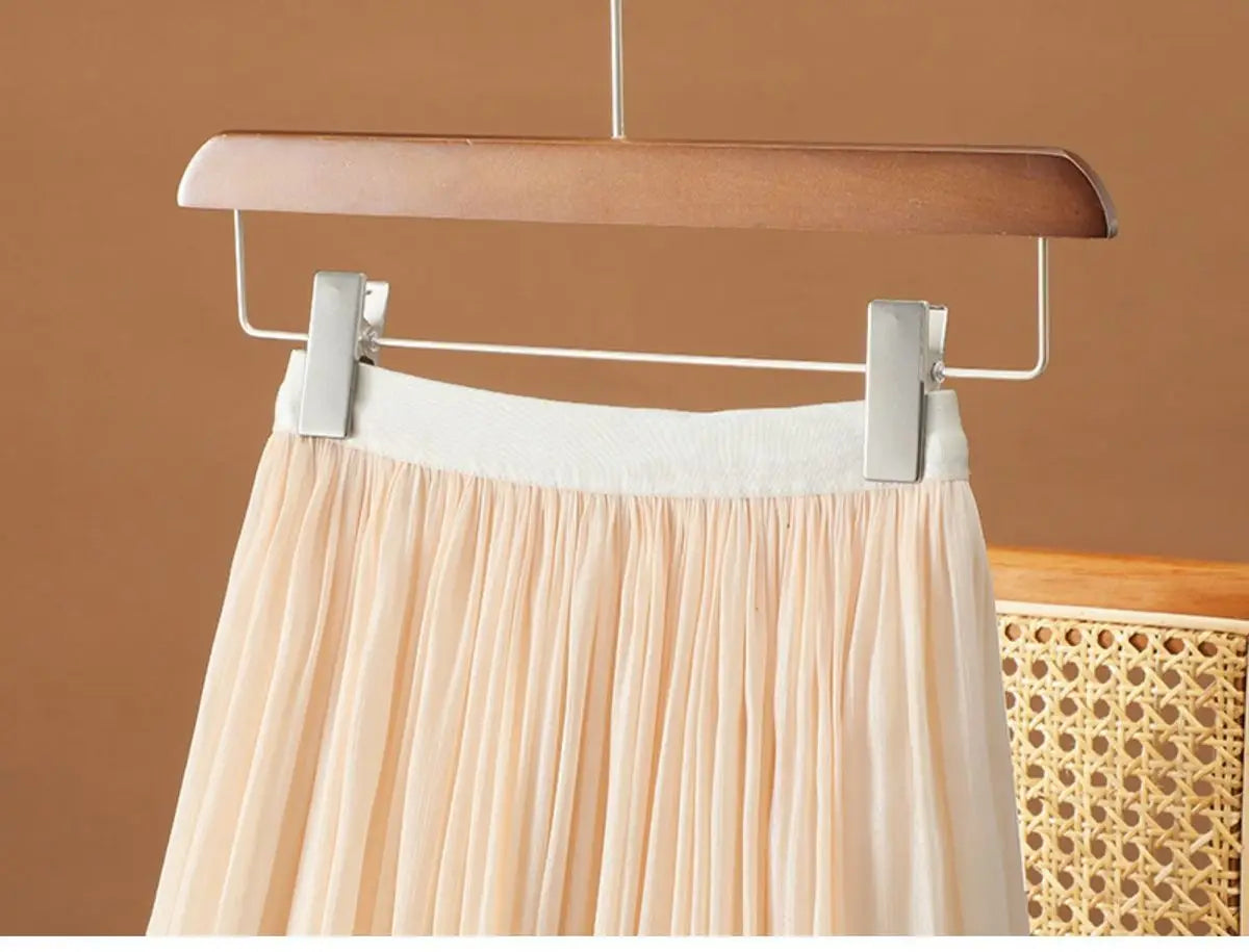 Ein sanftes pfirsichfarbenes, irrisierendes Chiffon-Faltenrock mit weißem elastischem Bund, der an einem Holzkleiderbügel hängt.