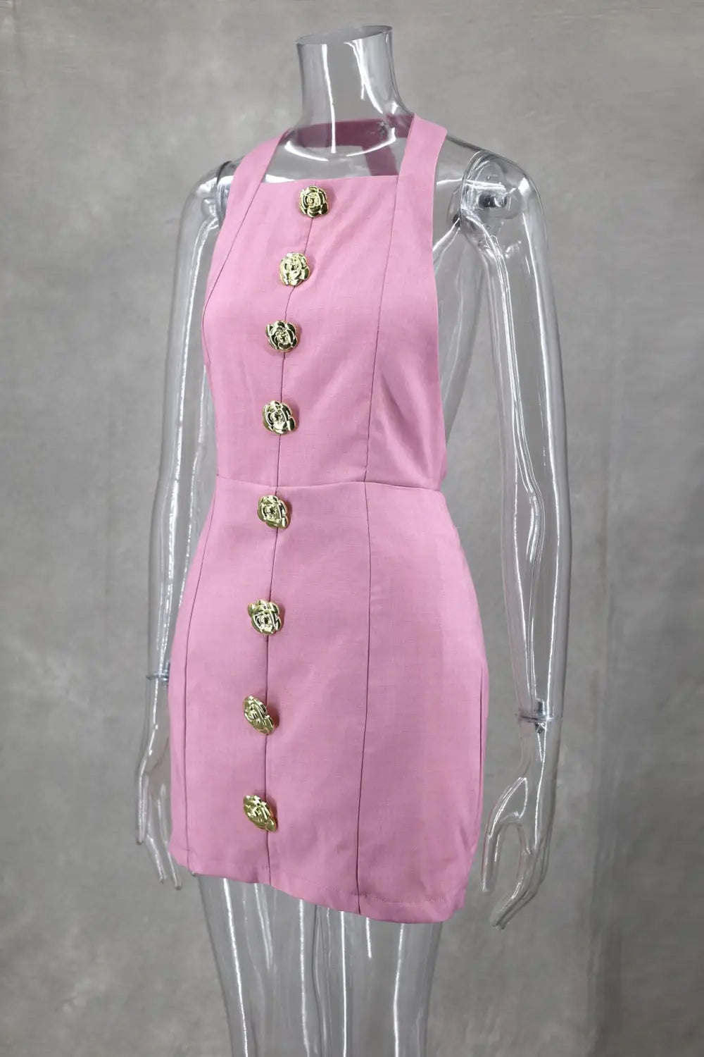 Une robe mini rose sans manches et dos nu avec des boutons décoratifs en forme de rose ornés d’un ton doré sur l’avant