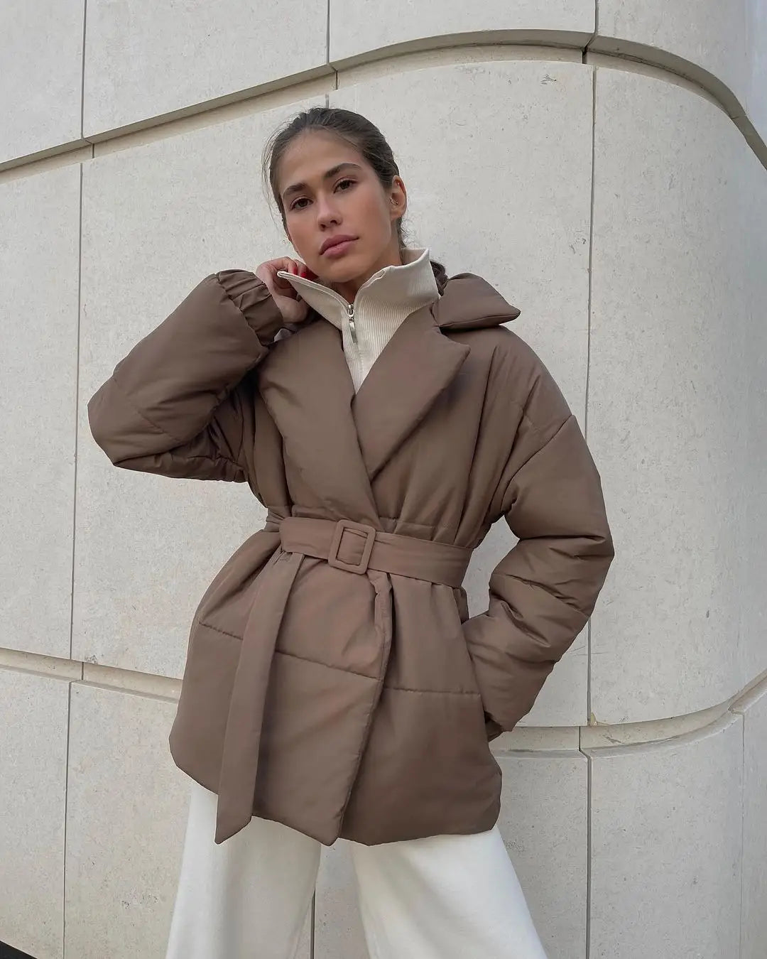 Eine gemütliche, taupefarbene gesteppte Pufferjacke für Damen mit Taillengürtel und hohem Kragen, für Winterbekleidung konzipiert.