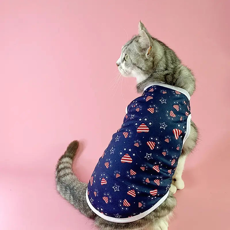 Un gato atigrado gris que lleva un chaleco de malla para perro con bandera estadounidense, camiseta para mascota en EE. UU., en azul marino con corazones a rayas rojo y blanco, estrellas y ribete blanco.