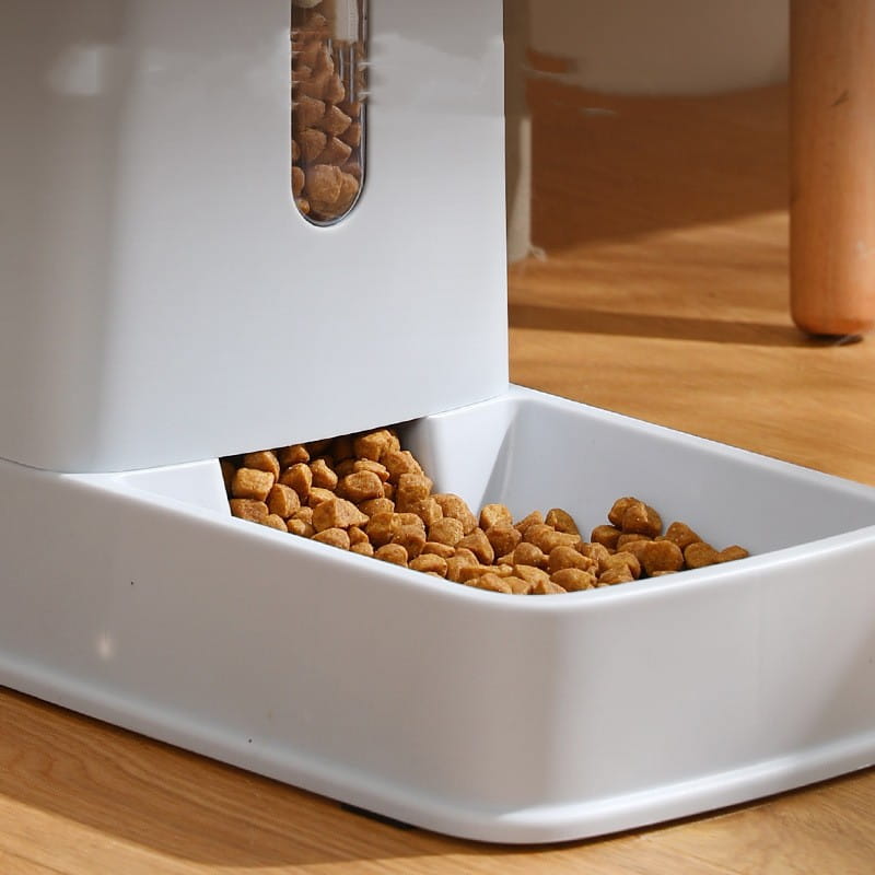 White plastic automatic pet feeder with clear window displaying stored kibble and tray filled with brown dry food pellets in an automatic pet feeder and water dispenser.