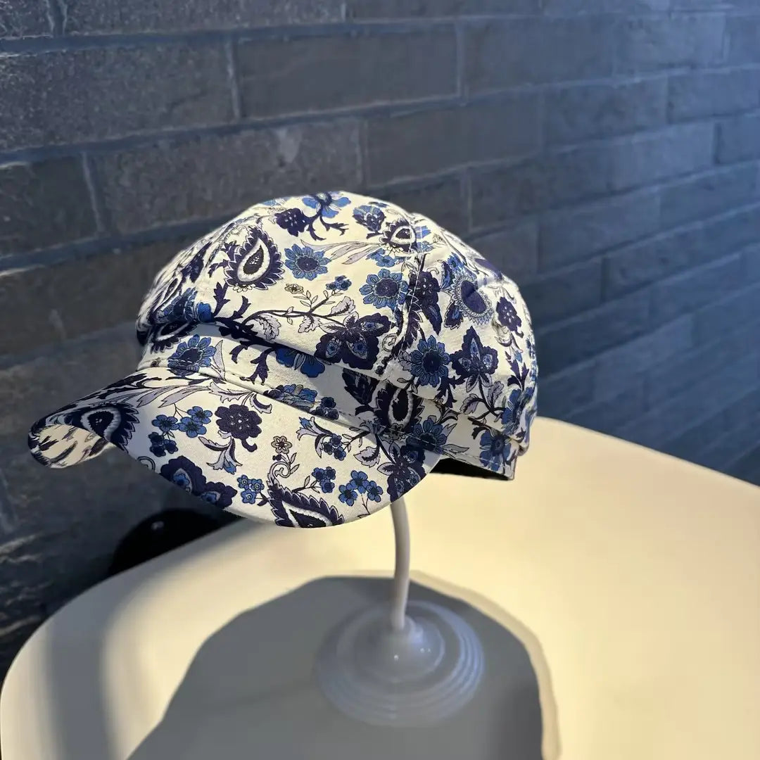 A floral newsboy cap for women with a white fabric base and intricate blue floral patterns featuring a classic flat brim design.