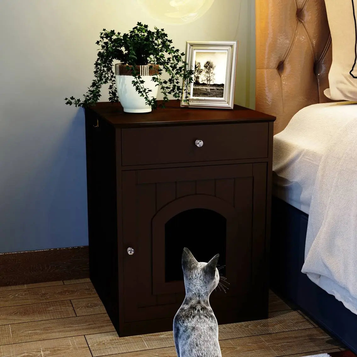 Eine Einfassung für eine Katzenkloschale aus dunkelbraunem Holz als Nachttisch mit Schublade und einer gebogenen Tür, oben ergänzt durch eine Topfpflanze und ein gerahmtes Foto.