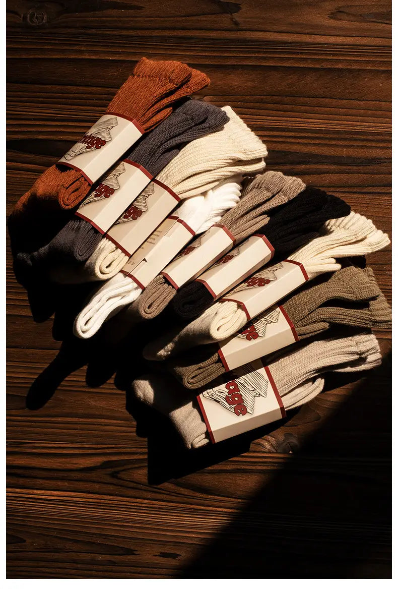 A stack of neatly folded vintage ribbed cotton long socks for men in earth tones including orange gray cream black and beige each wrapped with a branded paper band featuring red accents.