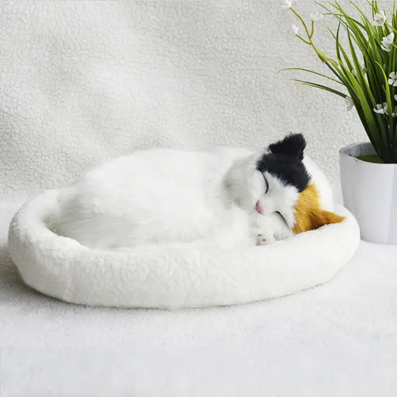 A plush white cat bed with a soft fluffy texture cradles a sleeping calico kitten in the realistic breathing plush cat figurine with pet bed.
