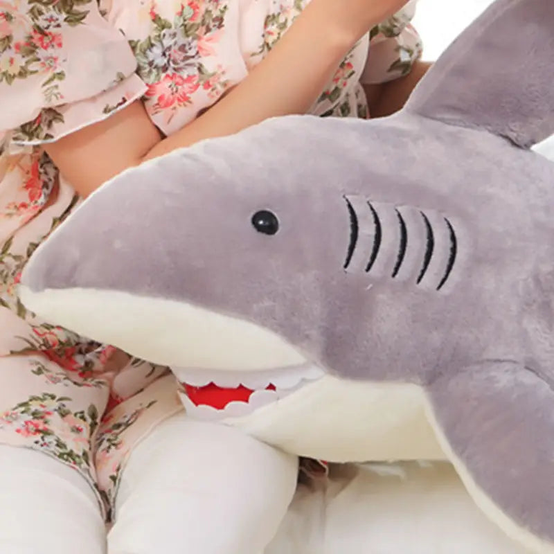 A plush gray shark pillow with white underbelly and red stitched teeth and gills designed as a soft toy.