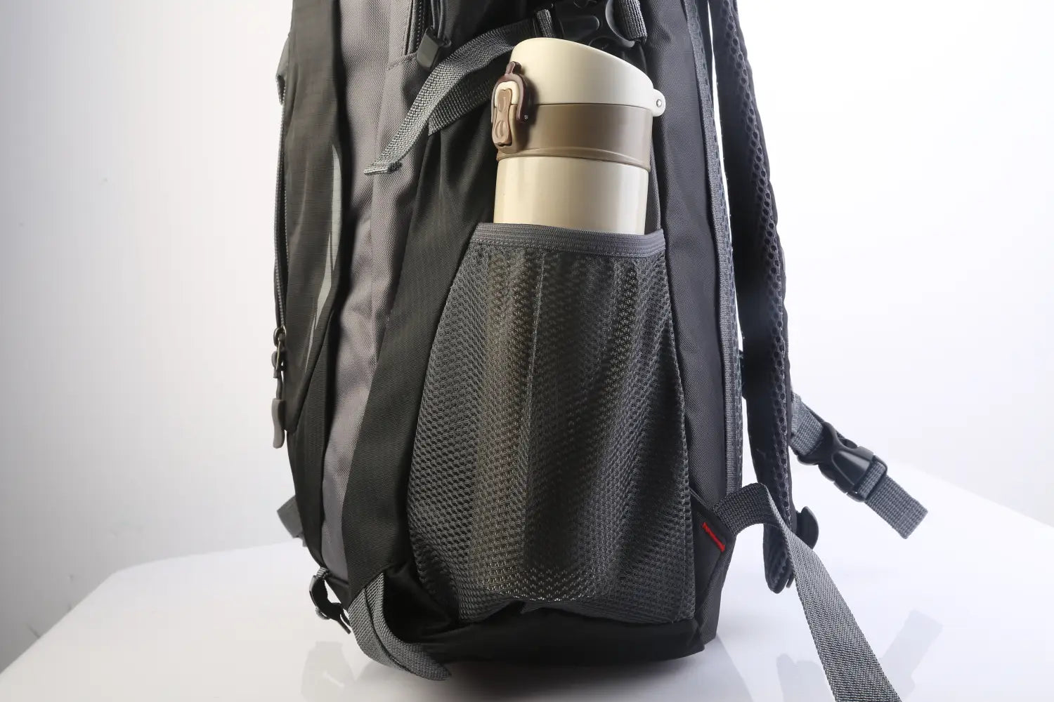 a beige and brown insulated travel mug with a metallic lid in the mesh side pocket of a black 36l lightweight waterproof hiking backpack with reflective stripes