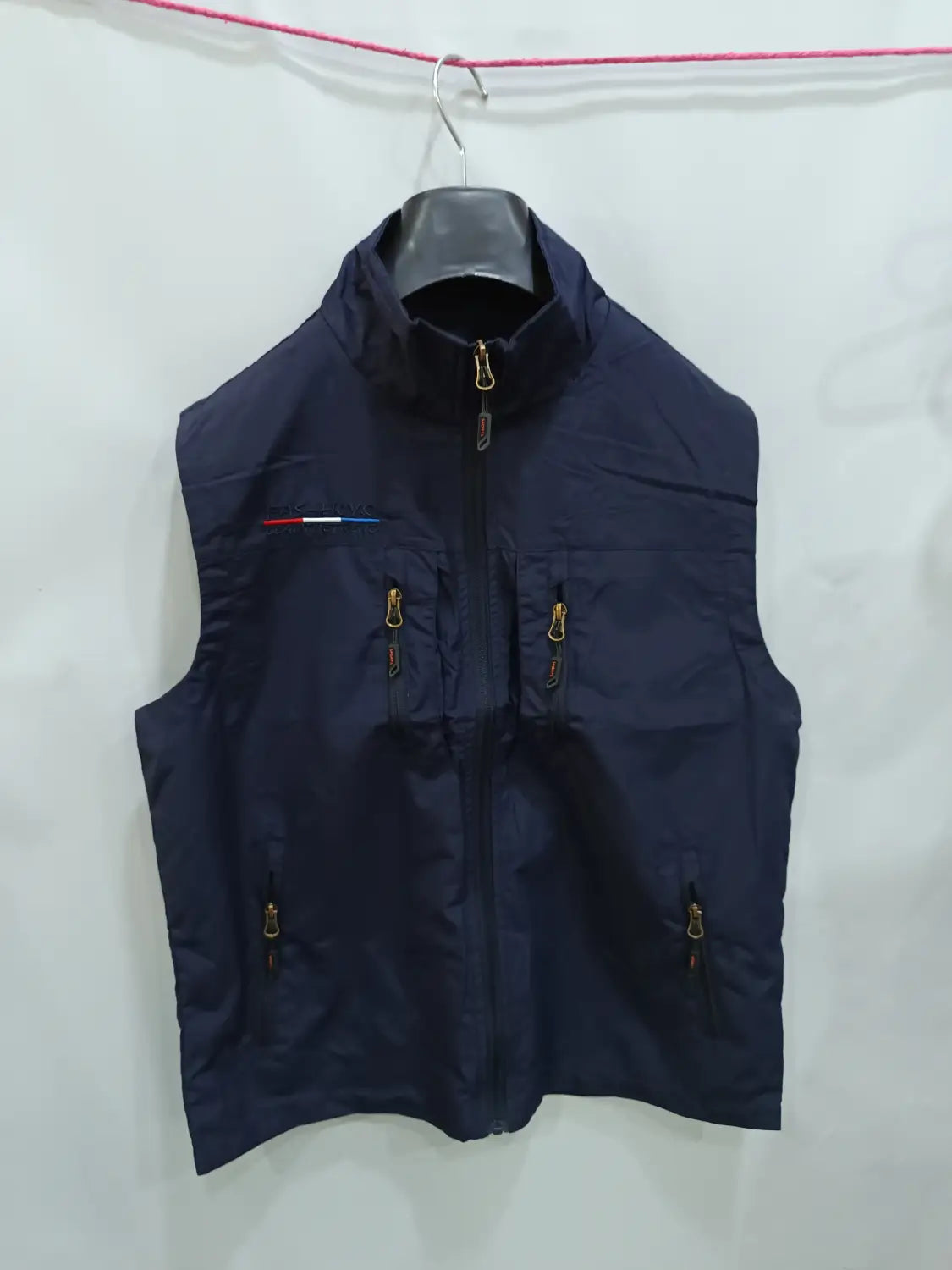 Chaleco utilitario sin mangas para hombre en azul marino oscuro con cremalleras de acabado dorado y un pequeño detalle de franja roja, blanca y azul en el lado izquierdo del pecho, con varios bolsillos y capucha.
