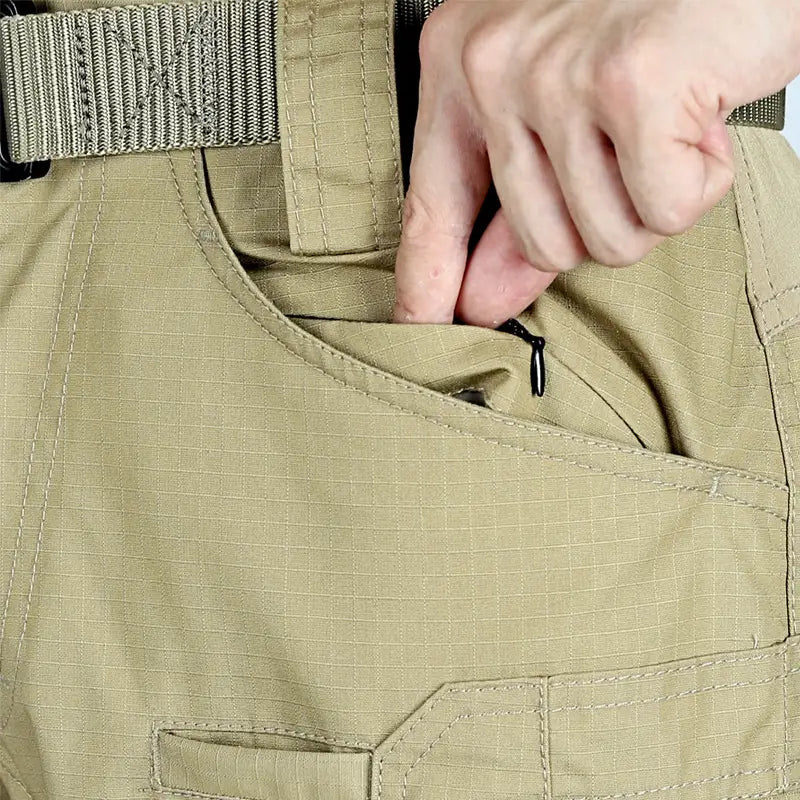 Pantalons tactiques P40 couleur kaki pour hommes : poches multiples, tissu à motif quadrillé, coutures renforcées, et main accédant à une poche zippée, conçus pour un usage militaire et en extérieur.