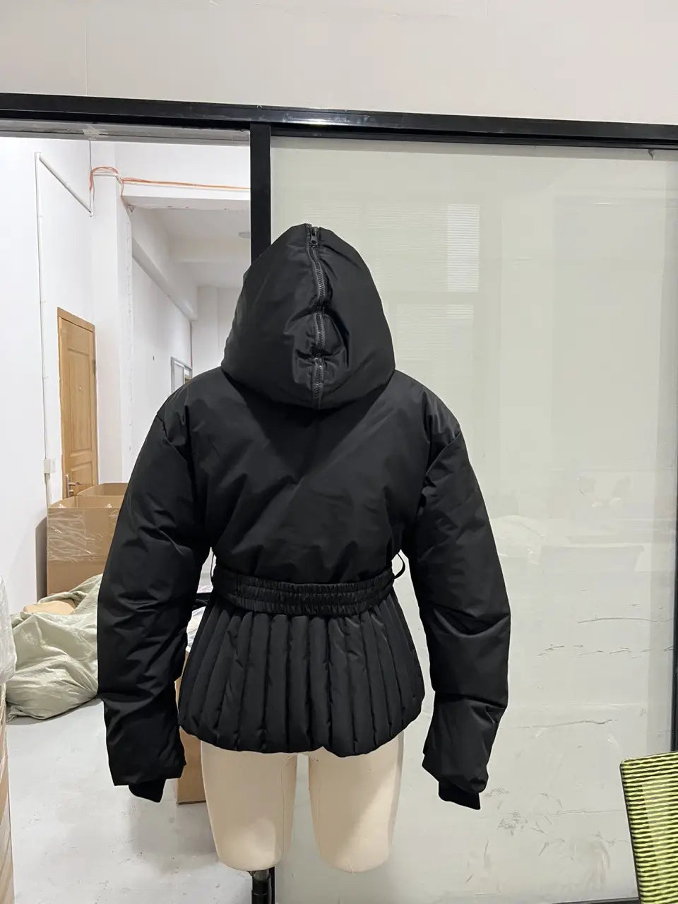 Women’s black belted hooded puffer coat featuring an elastic waistband and pleated skirt-like hem shown from the back.