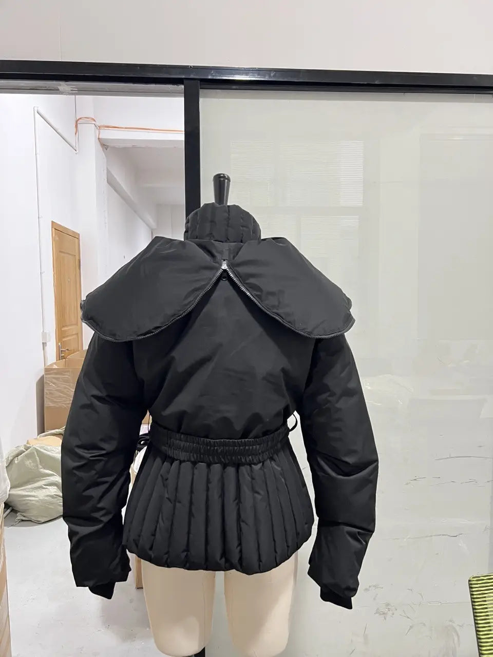 Black women’s belted hooded puffer coat featuring a wide structured collar and pleated skirt detail.