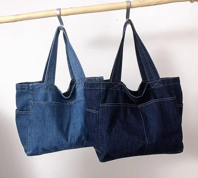Two denim tote bags in medium blue and dark indigo hang side by side from a wooden rod showcasing the large capacity Alma Latina shoulder bag.