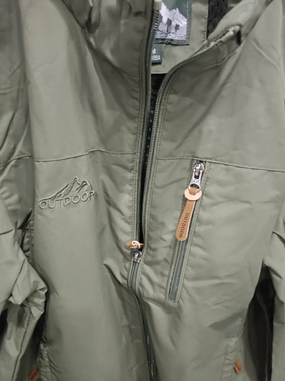 Chaqueta de senderismo con capucha impermeable color verde oliva, con logotipo de montaña bordado y tiradores de cremallera de cuero, diseñada para actividades al aire libre.