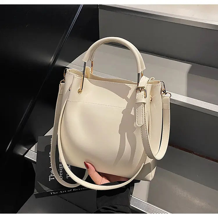 Cream-colored minimalist leather bucket bag with gold-tone hardware and a detachable shoulder strap.