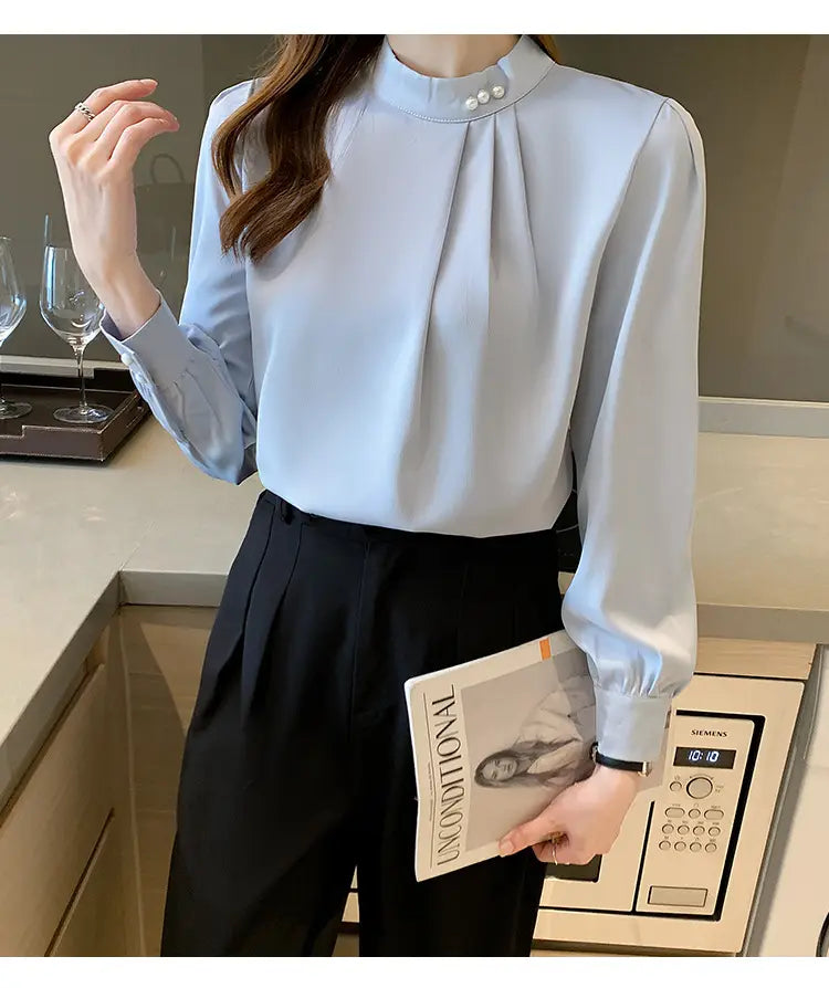 Blouse ample en satin bleu clair pour femmes, à encolure haute et trois accents de boutons évoquant la perle au niveau de l’encolure.