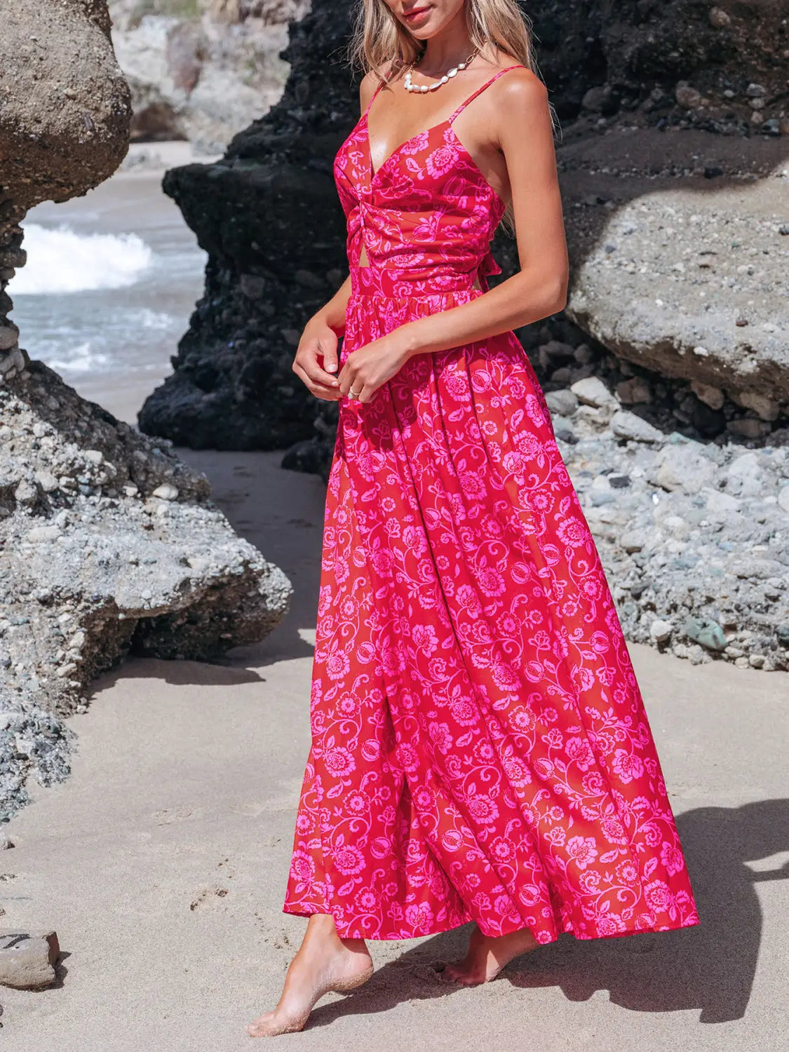 A vibrant pink floral knotted v neck spaghetti strap maxi dress with a fitted bodice flowing skirt and an elegant cutout detail at the waist.