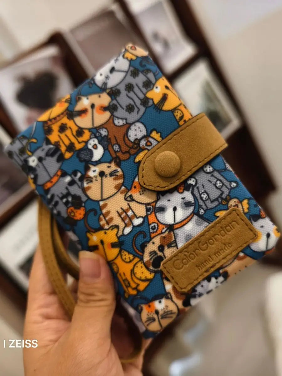 A blue fabric whimsical patterned canvas tri fold multi card wallet featuring a playful pattern of colorful cartoon cats and a tan leather-like flap closure with brand tag.