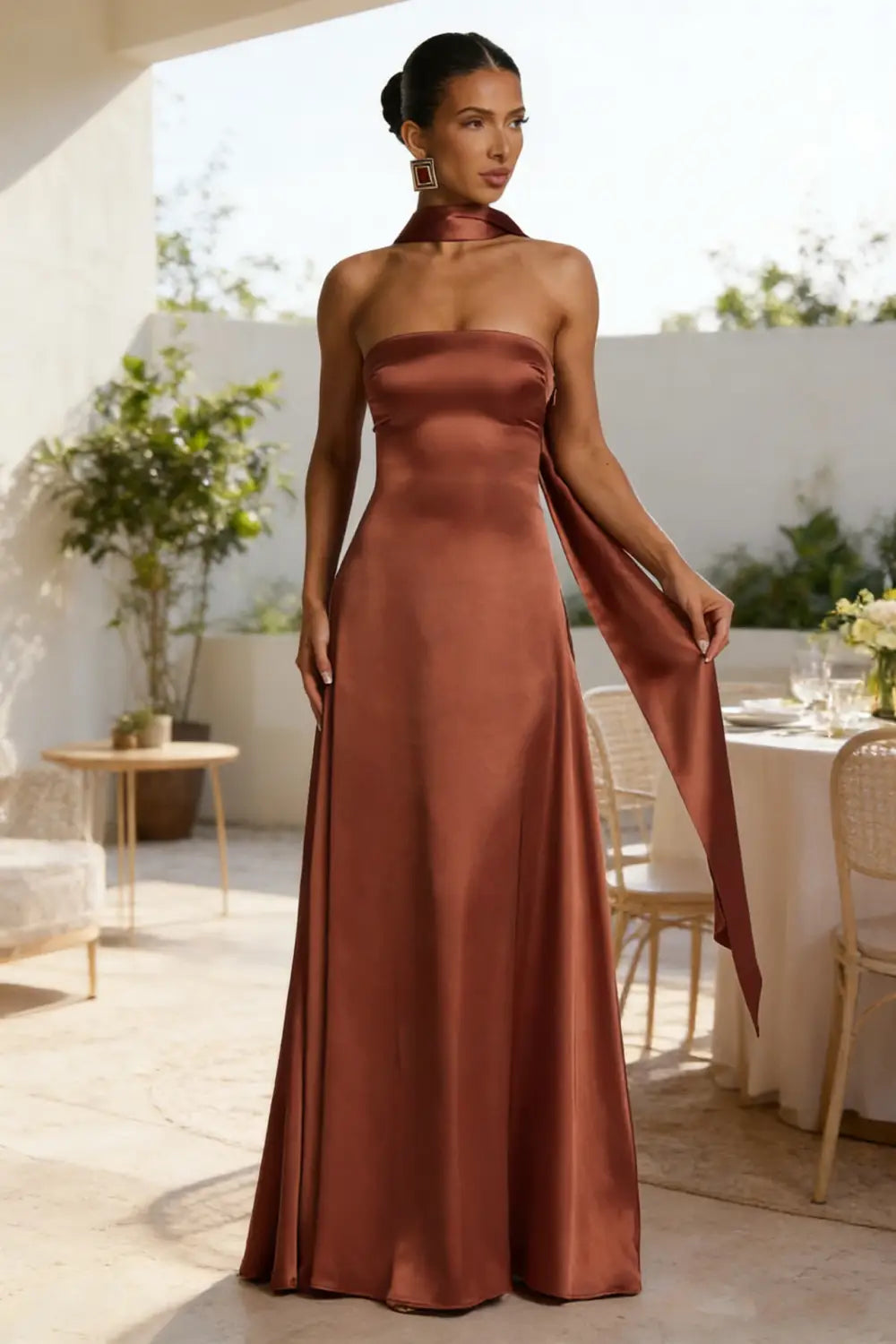 Un elegante vestido de noche sin tirantes de satén en color óxido, con un diseño fluido, escote tipo halter y detalle de tela drapeada.