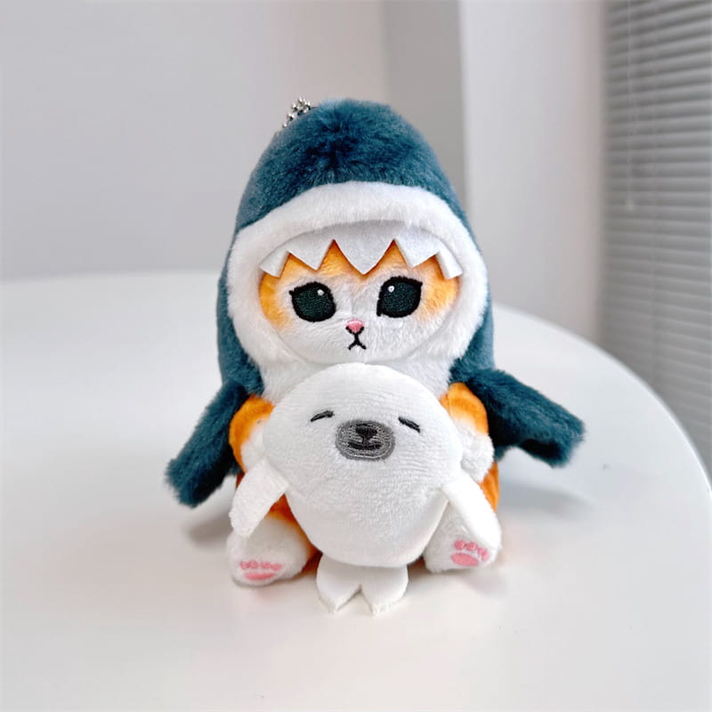 愛らしいキャリコ柄の猫のぬいぐるみキーホルダー。白い縁取りと鋭い歯のあるティール色のサメのフーディーを着用し、小さな白いアザラシのぬいぐるみを抱えています。日本の猫の人形（だるま）風のペンダント。