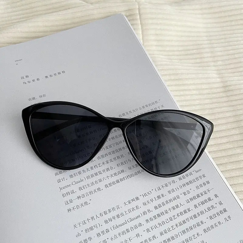 Lunettes de soleil œil-de-chat noires UV380 légères pour la mode, avec des verres gris foncé posées sur un livre ouvert.