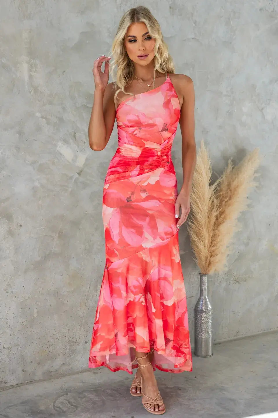 A coral and pink floral print one shoulder ruched asymmetrical maxi dress with a fitted bodice, ruched waistband, and flowing flared skirt.