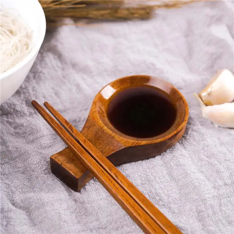 Un plato para salsa de inmersión de madera pulida con soporte para palillos, que incorpora un cuenco para sostener salsa de soja oscura con un acabado suave y brillante, y vetas naturales de la madera.