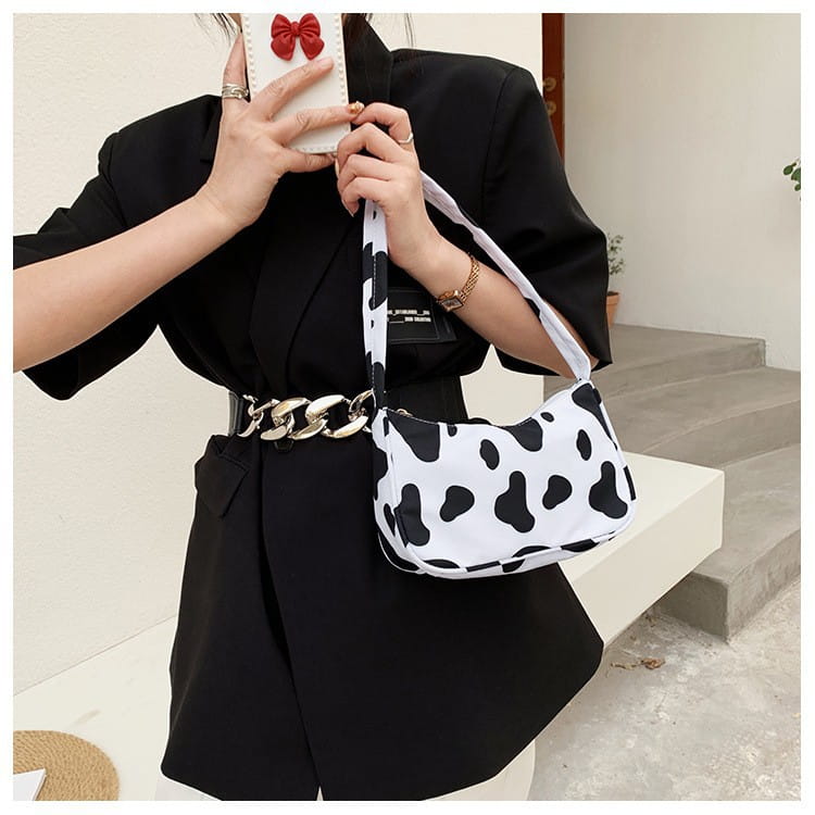 Sac bandoulière en toile blanche à imprimé vache, avec sangle assortie et ceinture en chaîne argentée, complétant la conception du sac sous-bras en nylon à imprimé léopard et à carreaux.