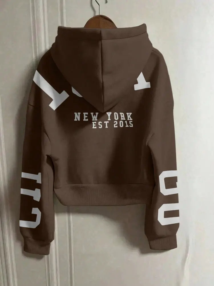 Sweat-shirt à capuche zippé marron court avec impression graphique, présentant le texte « New York Est 2015 » blanc et des motifs géométriques audacieux au dos et sur les manches, vu depuis le côté arrière