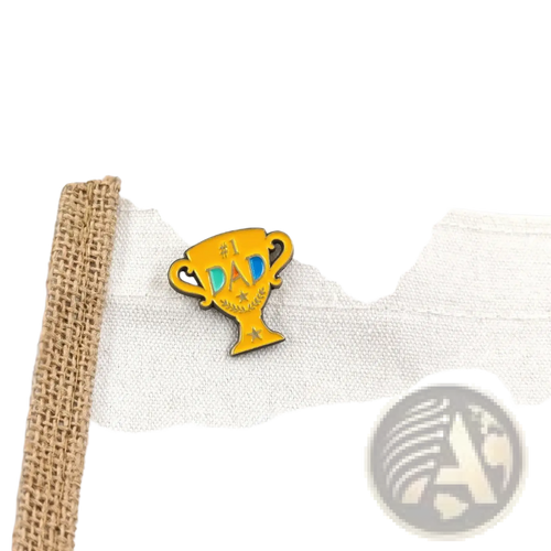 A vibrant yellow enamel pin shaped like a trophy with #1 dad text in blue and black and decorative stars