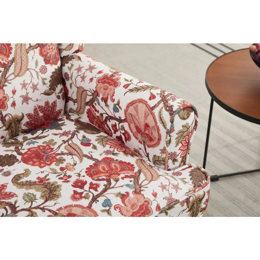 Una silla tipo ala con respaldo capitoné, tapizada en un tejido floral vibrante con flores llamativas rosadas rojas y marrones sobre un fondo blanco, con patas de madera retro.