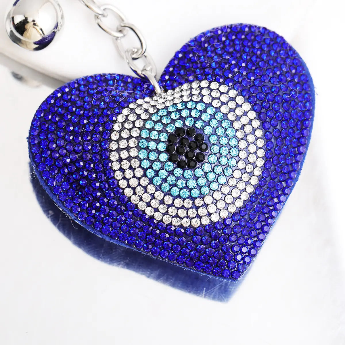 A heart-shaped keychain adorned with blue and silver rhinestones features a detailed evil eye design at its center and includes a blue rhinestone leather tassel.