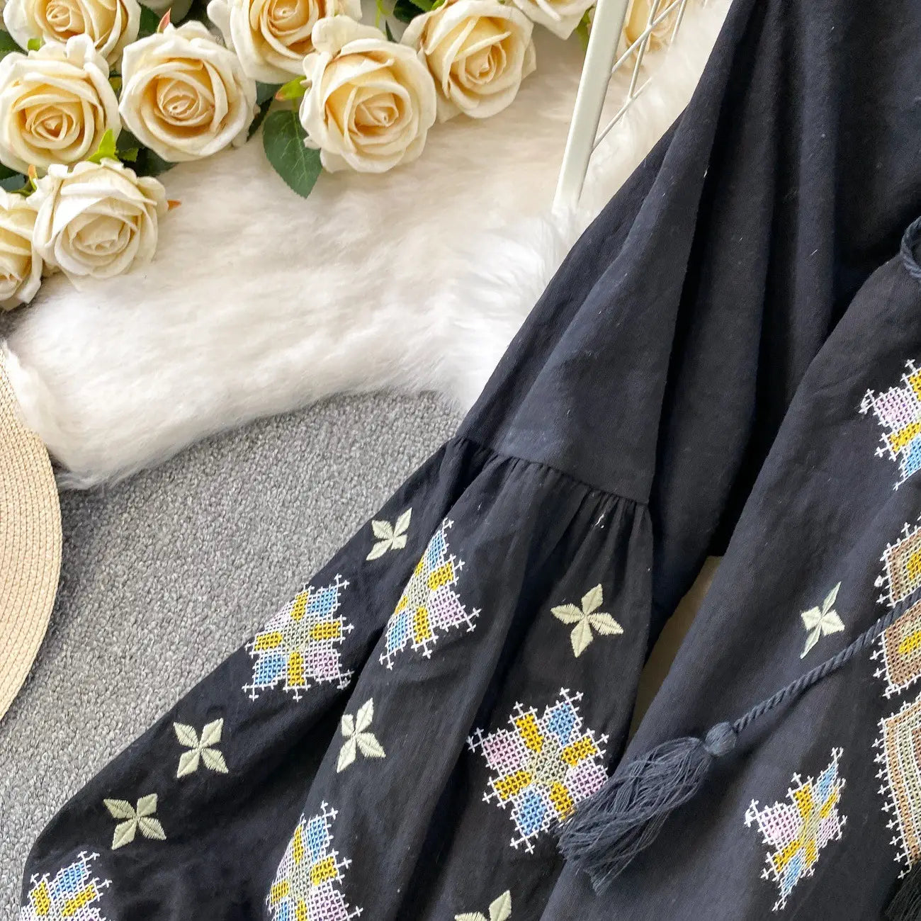 Black embroidered lantern sleeve tie neck blouse with colorful yellow blue and white cross-stitch patterns and tassel detail at the hem.