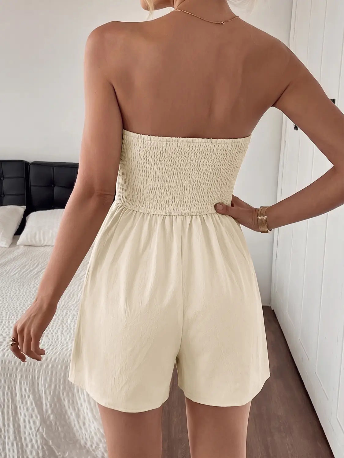 Cream-colored strapless pleated jumpsuit featuring a smocked bodice, fitted waistband, and loose-fitting shorts.