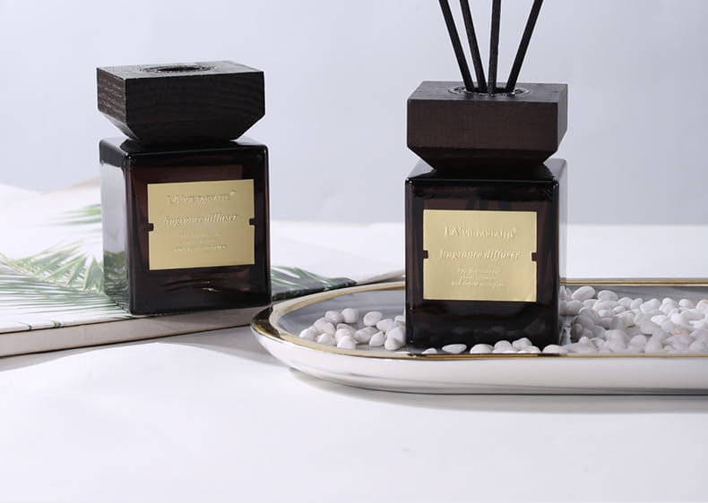 Deux diffuseurs à roseaux en verre noir pour parfum d’hôtel avec verre ambré foncé, dessus en bois noir et étiquettes de couleur dorée, l’un sur un magazine et l’autre dans un plateau blanc avec de petits galets.
