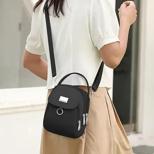 Schwarze kompakte Nylon-Umhängetasche mit Frontklappe, silberfarbenen Metallbeschlägen, Reißverschlüssen und Befestigungsmöglichkeit für ein Earbud-Etui.