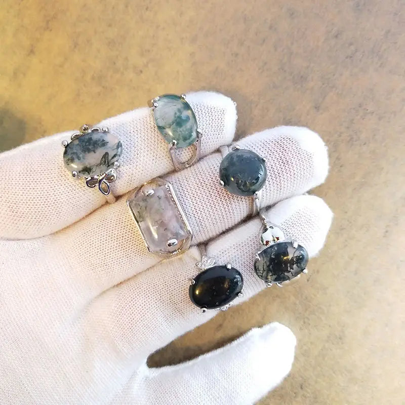 A hand in a white glove holds an assortment of silver tone statement rings featuring variously shaped and colored natural moss agate gemstones.