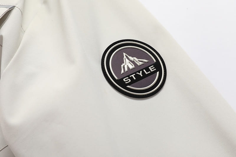 Ein runder schwarzer und grauer Patch mit einem Berglogo und dem eingestickten Wort „style“ ist auf dem weißen Stoff eines Herren-Wasserabweisenden Trenchcoats mit Kapuze angenäht
