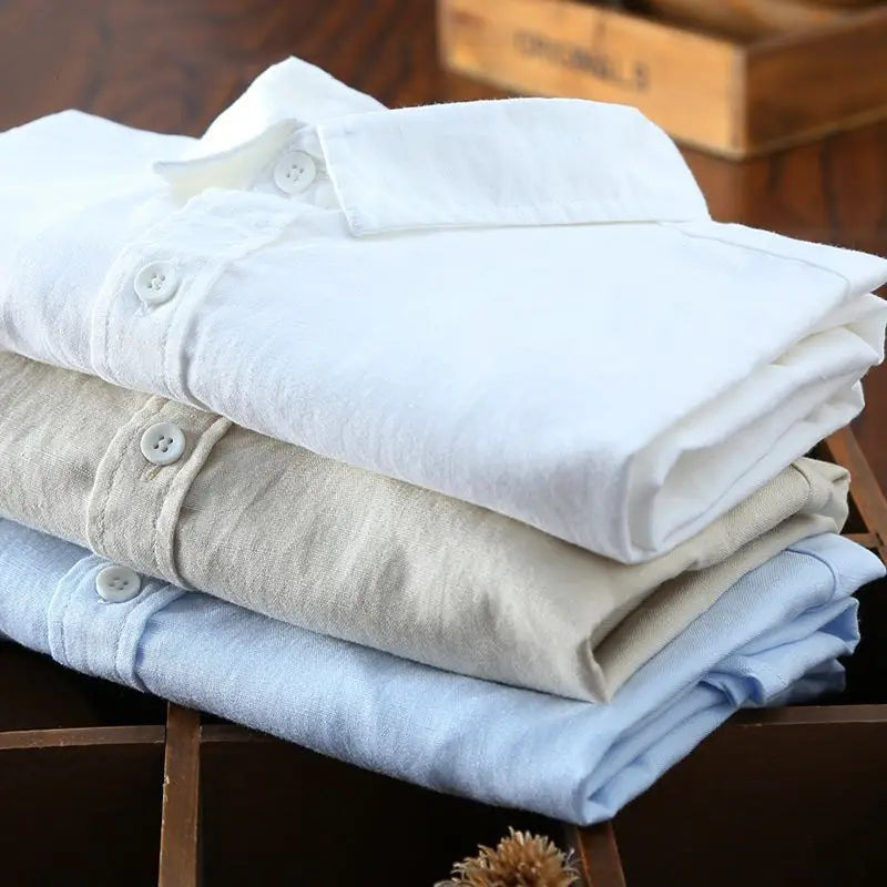 Ein Stapel aus drei ordentlich gefalteten Herren-Baumwoll-Leinen-Casual-Hemden mit langen Ärmeln und Knopfleiste in Weiß, Beige und Hellblau.
