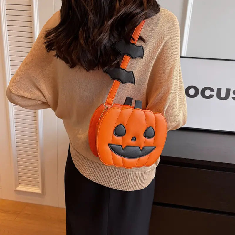 Orange pumpkin-shaped crossbody bag featuring black bat embellishments and stitched facial features with a detachable charm for festive use.