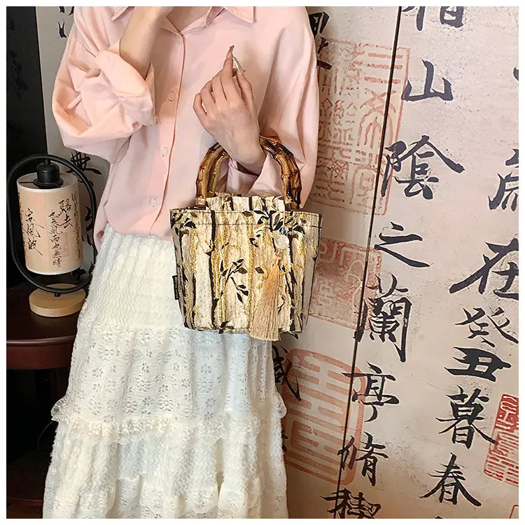 A woven bamboo-handled bucket bag with a gold and black floral forest pattern canvas featuring tassels at the bottom.