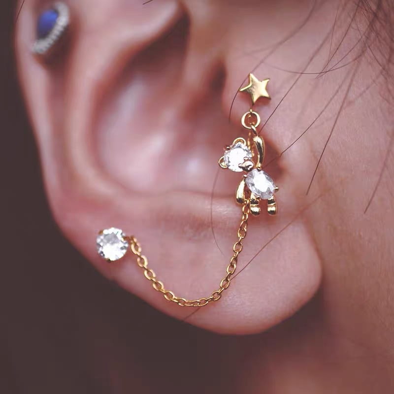 Boucle d’oreille asymétrique en strass à finition dorée, avec ours et pampilles en forme d’étoile, comportant un pendentif étoile, un élément décoratif en forme d’ours avec des pierres précieuses transparentes, ainsi qu’une chaîne délicate menant à un cristal rond.