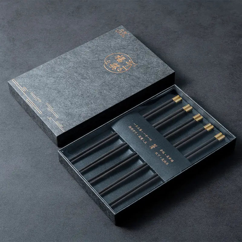 Schwarze hölzerne Essstäbchen mit goldfarbenen Spitzen, in einer eleganten dunkelgrauen Geschenkbox angeordnet, mit goldenen chinesischen Schriftzeichen, als Fünf-Paar-Set präsentiert.